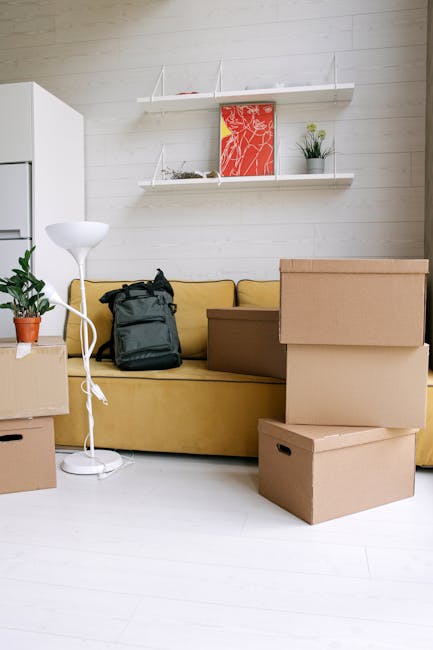 Inside a room with beige walls, a white sofa with crumpled fabric upholstery is positioned near cardboard moving boxes stacked against the wall. The boxes are sealed with packing tape and vary in size