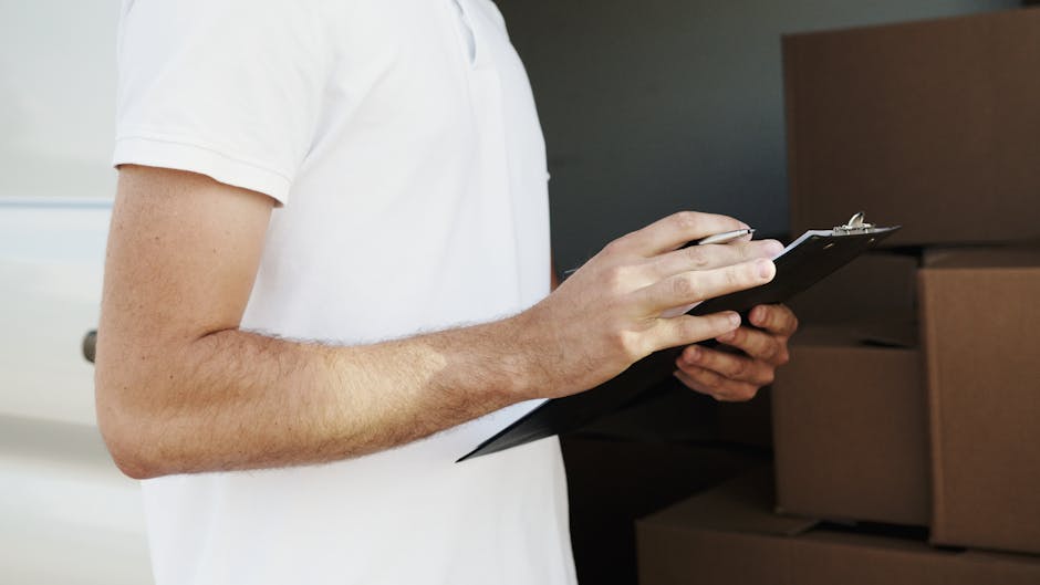 A woman wearing a white cap and black sleeveless top is outdoors, standing beside a stack of cardboard boxes. She holds a pen and a clipboard, appearing to take notes or inventory during a house remov