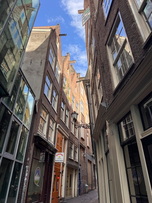 A narrow urban alleyway surrounded by tall, multi-storey brick buildings with large windows, some with white frames and others with black, under a partly cloudy blue sky. On the right, a white-painted building with a decorative sill and a small sign is visible. On the left, modern glass-fronted buildings reflect the sky and neighboring structures. An ornate black street lamp is mounted on the right building, extending horizontally over the alley. The alley's surface is paved with cobblestones, and the scene appears to be in a city centre, typical of an area where home relocation or furniture transport services, such as those provided by Man and Van Clapton, might operate in narrow streets.