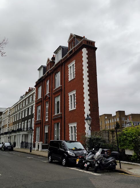 A tall, narrow red brick building with white decorative accents along its corners and window frames, situated on a residential street in Clapton. The building features several upper-story windows, with some open or covered, and a small dormer window at the roof level. The street below shows parked black cars and two motorcycles, with a typical London-style lamp post nearby. The sky is overcast with grey clouds, and neighboring buildings with similar Victorian architecture are visible on either side, indicating a dense city environment suitable for home relocations or furniture transport. This scene relates to house removals and moving services, highlighting the urban setting for property transfer operations.
