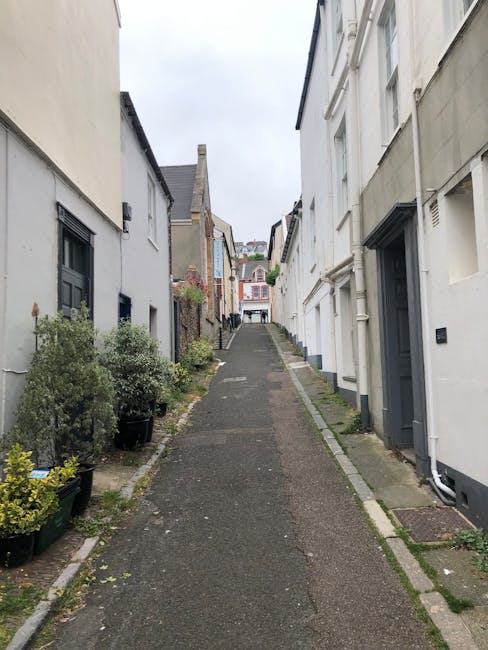 A narrow urban alleyway surrounded by tall, multi-storey brick buildings with large windows, some with white frames and others with black, under a partly cloudy blue sky. On the right, a white-painted building with a decorative sill and a small sign is visible. On the left, modern glass-fronted buildings reflect the sky and neighboring structures. An ornate black street lamp is mounted on the right building, extending horizontally over the alley. The alley's surface is paved with cobblestones, and the scene appears to be in a city centre, typical of an area where home relocation or furniture transport services, such as those provided by Man and Van Clapton, might operate in narrow streets.