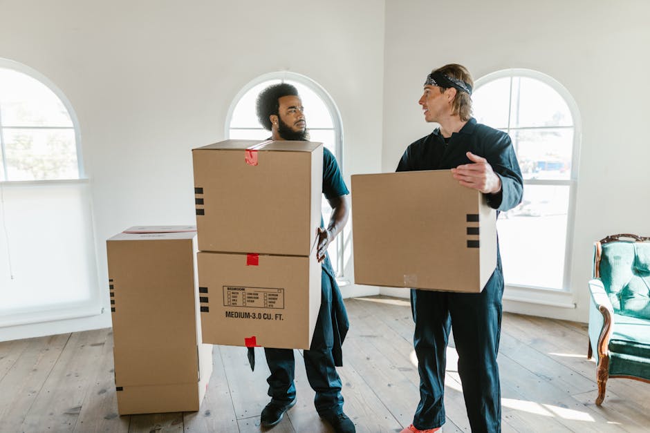 A young woman sitting cross-legged on a wooden floor inside a room, surrounded by numerous cardboard boxes used for packing during a house move. She is inside an open box and is playfully tossing pack