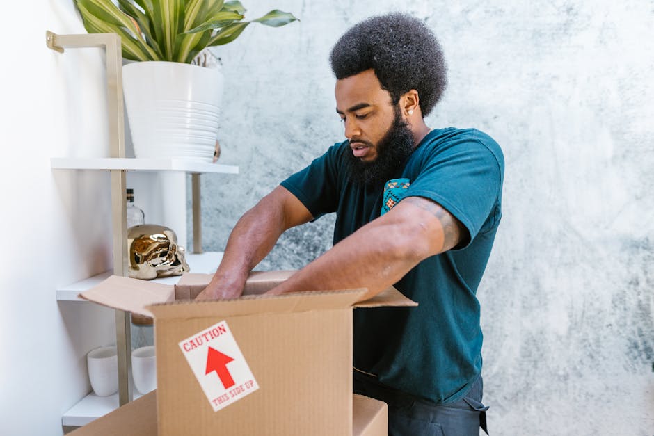 A man with a curly afro hairstyle and a beard is standing inside a home, packing belongings into a cardboard box marked with a red 'CAUTION' label and an upward arrow indicating 'this side up.' He is wearing a dark blue T-shirt and is focused on carefully placing items into the box, with both hands inside. The scene is set against a light textured wall, with a white shelving unit nearby holding a large potted plant, a decorative golden skull, and white ceramic cups. Natural lighting illuminates the area, emphasizing the packing process involved in home relocation or furniture transport, which is typical for professional removals services such as those provided by Man and Van Clapton. This image illustrates the careful packing stage in the moving process, emphasizing handling fragile or important items securely during house removals.
