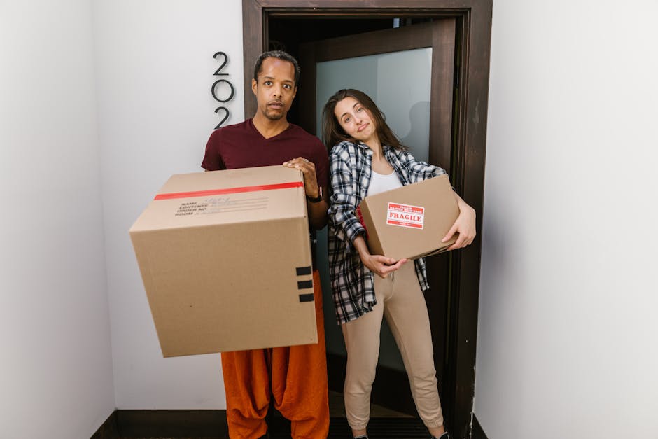 A man and a woman stand inside a doorway in a property, preparing for a home relocation. The man is wearing an orange and brown outfit and holds a large cardboard box with black tape at the top, while the woman is dressed in a checkered shirt and light-colored trousers, carrying a smaller cardboard box labeled 'Fragile' with red and white packing tape. Behind them, the door frame and part of the interior wall are visible, with a slightly open door revealing a glimpse of a room. The lighting is bright and natural, indicating daytime. The scene captures the packing and moving process, with both individuals focused on transporting boxes, demonstrating the steps involved in furniture transport and packing as part of professional removals services, such as those offered by Man and Van Clapton.