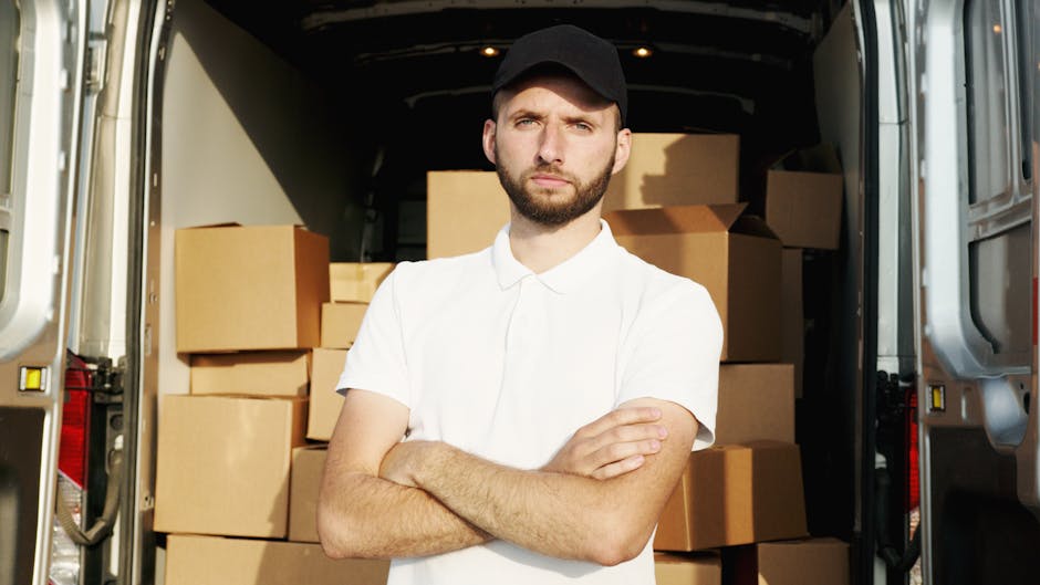 A man with a beard and short dark hair, wearing a black cap and a white polo shirt, stands inside the open back of a van loaded with neatly stacked cardboard boxes of various sizes, some wrapped in pl